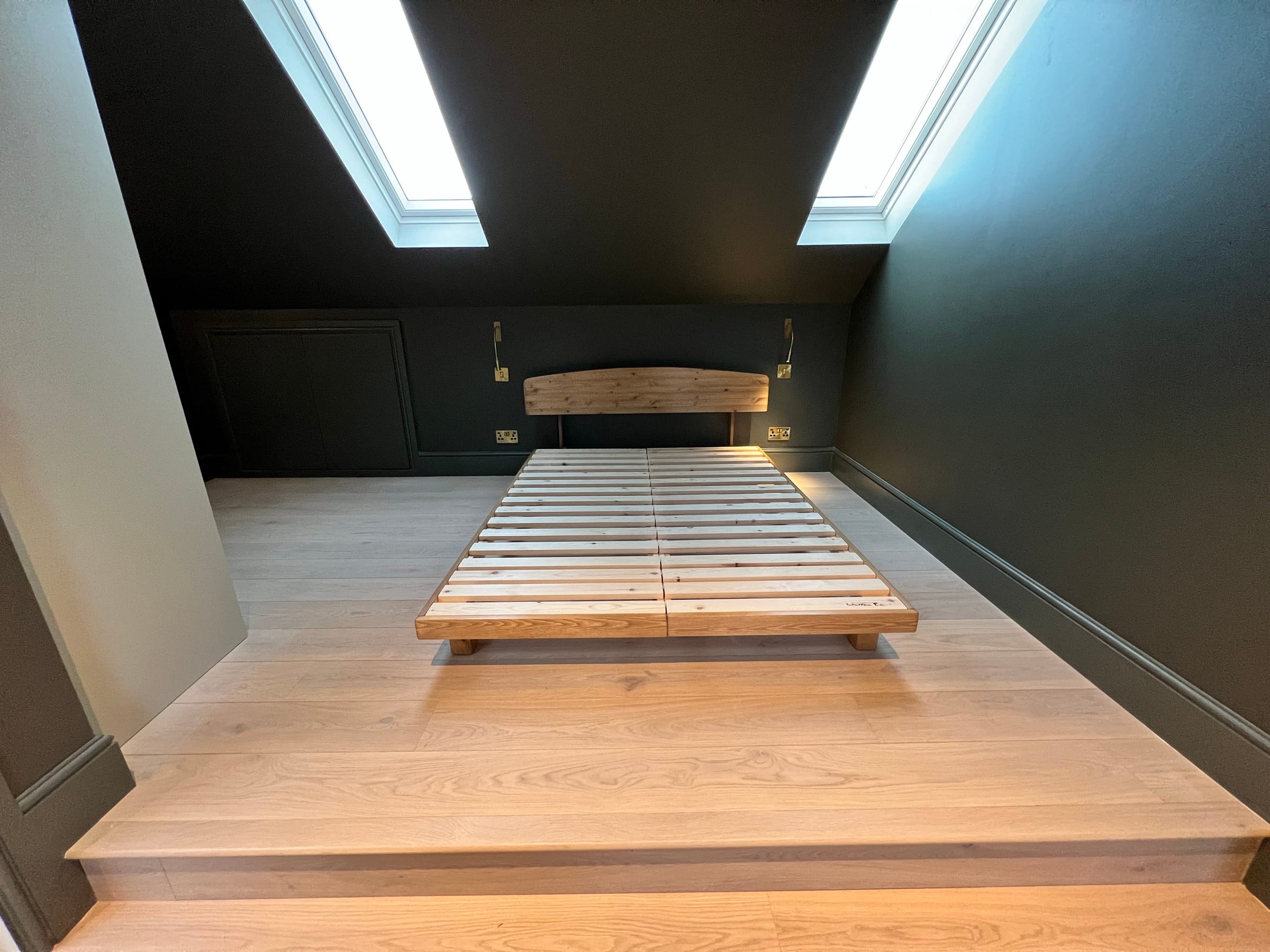 A completed loft conversion bedroom in dark green with an oak platform bed and a rooflight set into the slope, by 2VP