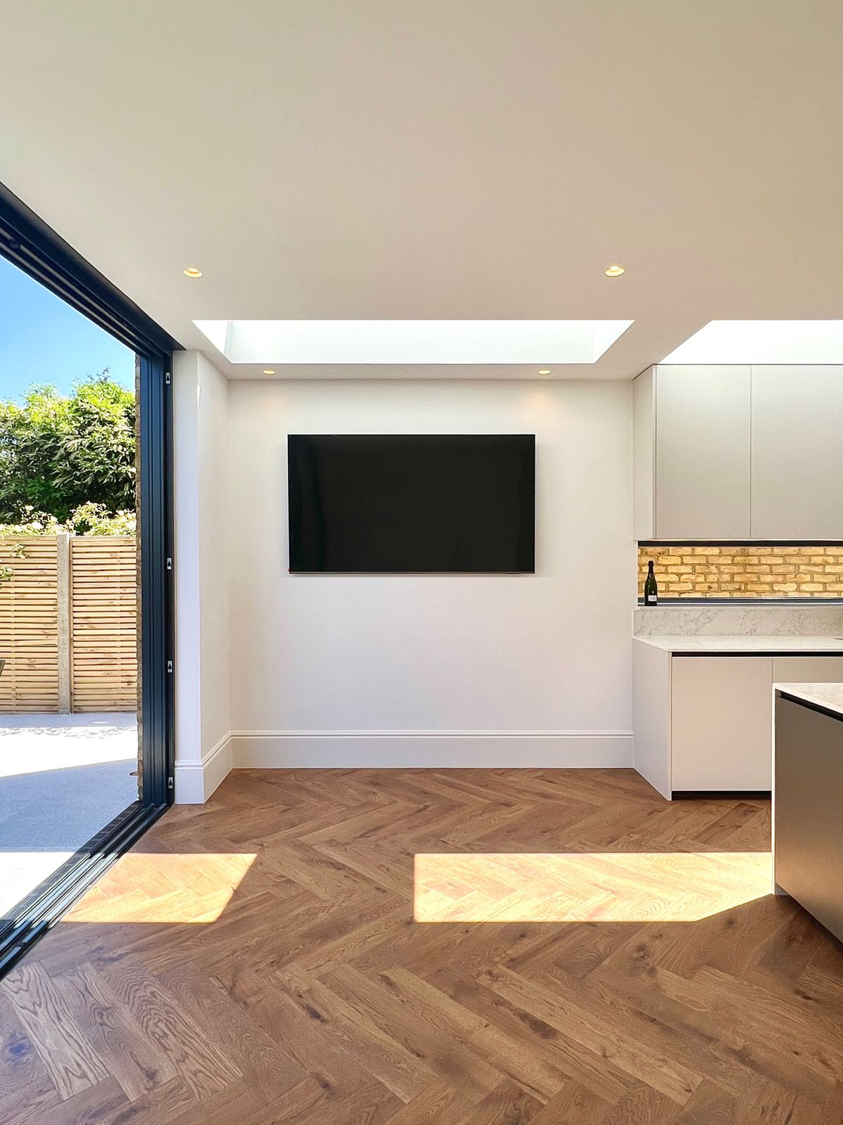 Handleless kitchen opening to the garden — Marble worktop, brick splashback lit by a warm strip light, herringbone oak floor, bifolds to the garden