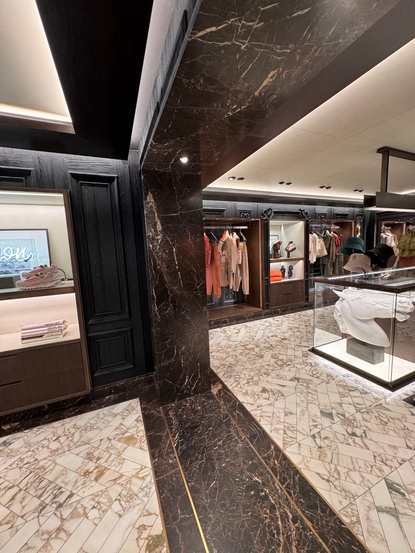 Marble-clad column and chevron floor — Moncler · London · Emperador borders, brass inlay, stained oak panelling