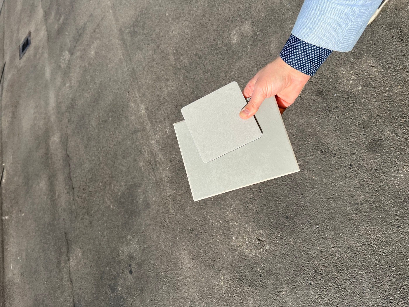 A hand holding two small pale resin sample panels up against a grey resin floor outdoors